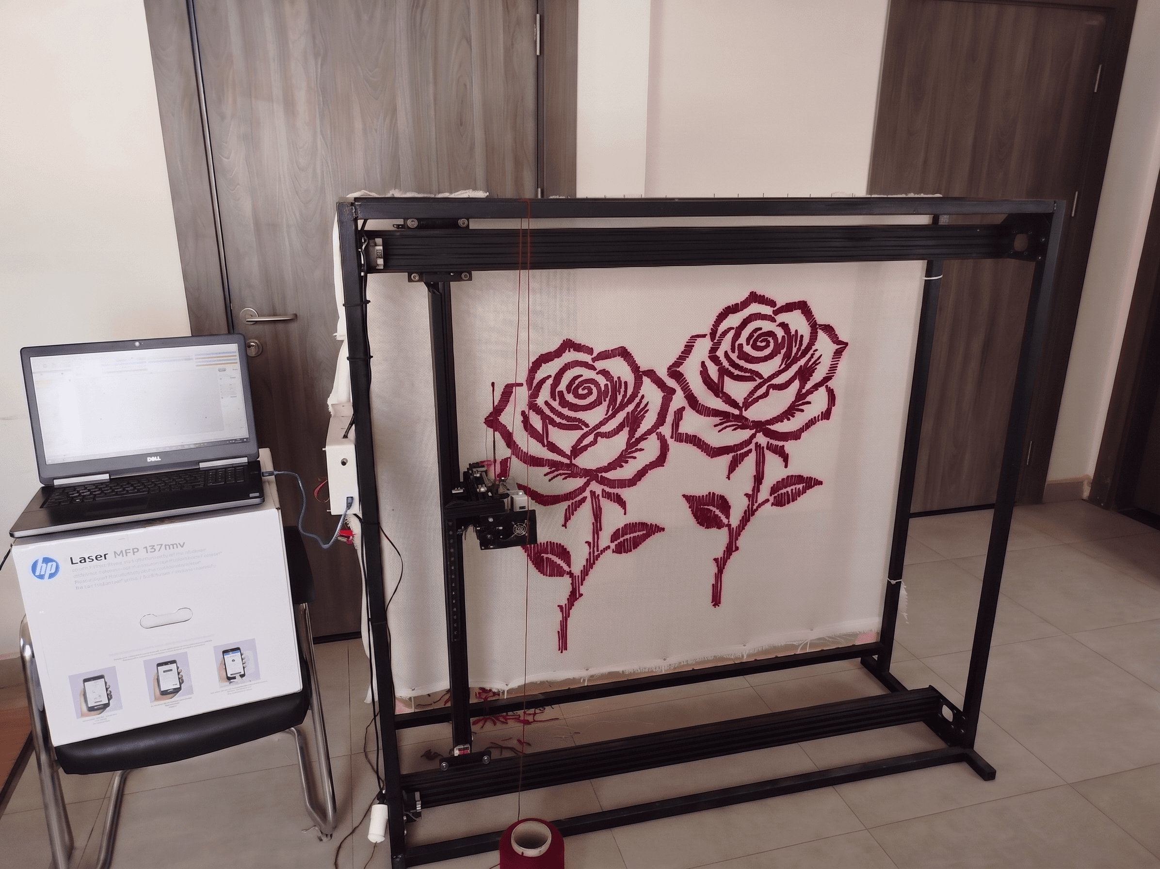 Large CNC machine tufting two red roses on white fabric, controlled by a nearby laptop.