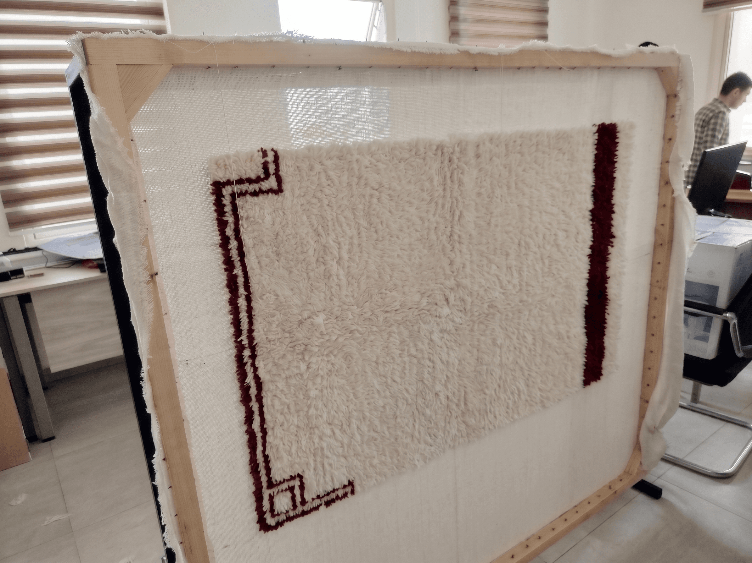 White shaggy rug with maroon borders stretched on a large wooden frame for tufting.