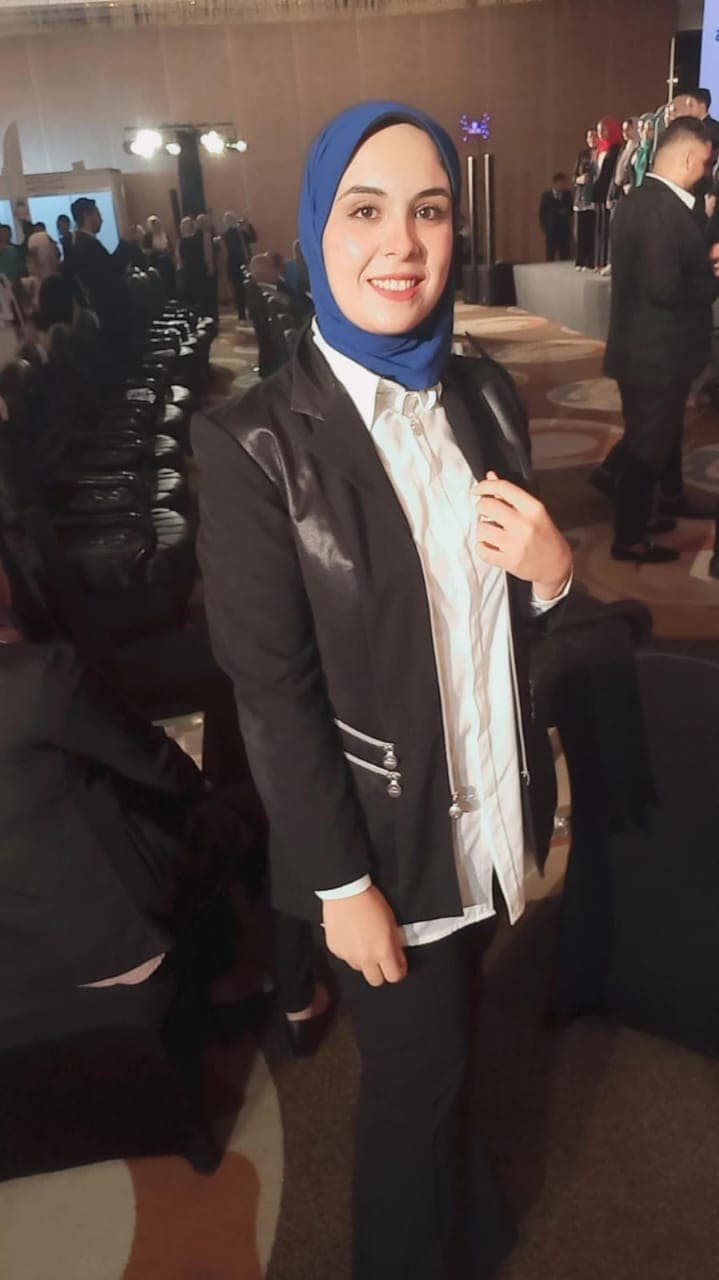 Woman in a blue hijab and black blazer smiling at a formal indoor event.
