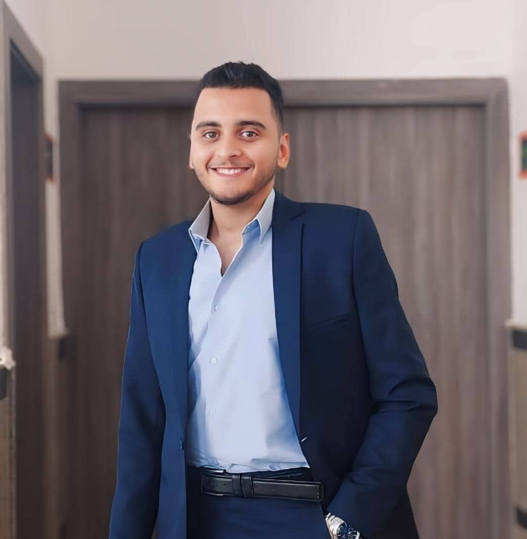 Smiling young man wearing a navy blue suit and light blue shirt standing indoors.