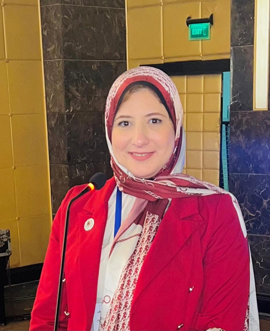 Woman in a red blazer and patterned hijab smiling while standing behind a microphone.