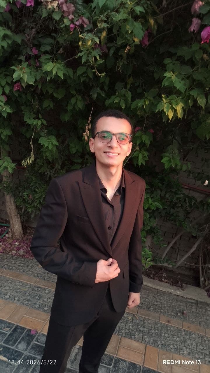 Smiling man in a dark suit and glasses stands before lush green garden foliage.